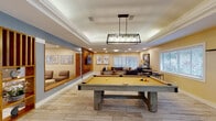 This single-floor community facility is thoughtfully designed to accommodate a variety of social and recreational activities. The space is unified by light wood-look tile flooring throughout, warm cream and soft blue accent walls, and a combination of recessed and pendant lighting that creates a bright, welcoming atmosphere.

The centerpiece of the recreational area is a full-size billiards table set beneath a modern rectangular pendant light fixture with a tray ceiling overhead. Adjacent to the game area is a spacious lounge featuring a large sectional sofa, woven accent chairs, and coastal-inspired artwork, all arranged around a flat-screen TV mounted on a warm wood slatted partition wall.

An open gathering area connects to a stylishly appointed kitchen equipped with a large island with bar seating, dark cabinetry, subway tile backsplash, stainless steel appliances, and pendant lighting. A casual dining area with a round table and woven chairs sits nearby, making the space ideal for community events.

A flexible multipurpose room features a tray ceiling, interior service window, and access to utility areas, while a separate bright room with French doors opening to the outdoors provides a casual seating nook and water station. Natural light pours in through multiple windows fitted with plantation shutters, enhancing the airy, coastal ambiance throughout the entire facility.