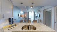 This contemporary single-floor residence showcases a thoughtfully designed open-plan layout that seamlessly connects the kitchen, dining, and living areas. The kitchen features a generous granite-topped island with a double stainless steel undermount sink, serving as both a functional workspace and a natural dividing point between the cooking and living zones. Sleek pendant lights hang above the island, casting warm illumination over the stone countertops.

The exposed concrete ceiling adds an industrial-chic character to the space, contrasting beautifully with the soft blue-grey walls that lend a calming, modern aesthetic throughout. The living area is comfortably appointed and benefits from abundant natural light streaming in through a floor-to-ceiling glass sliding door that leads to a private balcony, offering views of the surrounding cityscape.

Decorative wall accents, including a sunburst mirror and sculptural wall art, add personality and visual interest to the space. The dining area sits between the kitchen and living room, creating a natural flow for everyday living and entertaining. Overall, the residence strikes a balance between urban sophistication and comfortable, livable design.