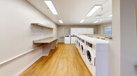 Located at 1175 Lake Blvd in Davis, California, this communal laundry facility offers a clean, functional, and well-organized space designed to serve its users efficiently. The single-floor layout is thoughtfully arranged to maximize usability and flow.

The main laundry area is lined with a full row of front-loading washing machines and stacked dryers along one wall, providing ample capacity for residents. A long folding counter with an overhead shelf runs along the opposite wall, offering generous workspace for sorting and folding laundry. Warm wood-look flooring spans the entire space, contributing to a welcoming atmosphere.

Natural light enters through multiple windows, complemented by flush-mounted fluorescent ceiling panels and a ceiling fan that help keep the room bright and comfortable. A utility sink with white cabinetry is conveniently positioned within the space for hand-washing or other needs.

A secondary entry area features warm beige-toned walls, additional wood-look flooring, and a small bookshelf with reading materials — a thoughtful touch for those waiting. A community bulletin board near the entrance displays announcements and local information, reinforcing the communal nature of the facility. Overall, the space is clean, practical, and well-maintained, making it a reliable amenity for daily use.