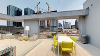 This sophisticated rooftop terrace is designed for elevated urban living, offering sweeping panoramic views of a dynamic city skyline. The space is thoughtfully divided into distinct functional zones, each finished with large-format light stone tile flooring that lends a clean, contemporary aesthetic throughout.

A central outdoor kitchen serves as a focal point, constructed with striking granite countertops and equipped with a sink, providing an ideal setup for al fresco entertaining. Teal wire-frame bar stools line the counter, adding a modern, colorful accent. Adjacent to the kitchen, a dedicated dining area features a crisp white table surrounded by vibrant chartreuse chairs, creating a bold pop of color against the neutral palette.

A separate lounge area invites relaxation with plush, neutral-toned sectional seating accented by teal and geometric-patterned throw pillows, complemented by sculptural planters with agave and succulents. A sleek floor lamp adds ambiance to this retreat zone.

Glass panel railings encircle the terrace, preserving unobstructed sightlines of the surrounding cityscape. A partially covered corridor with full-height glass walls connects the terrace to the building's interior, maintaining visual continuity between indoor and outdoor spaces. The overall design balances modern architecture with vibrant furnishings, creating an inviting and functional rooftop destination.