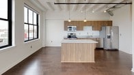 This two-bedroom, two-bathroom unit at Checker showcases a distinct industrial loft aesthetic, blending modern finishes with architectural character throughout. Soaring ceilings with exposed white-painted structural beams and visible ductwork create a spacious, open ambiance across the single-floor layout. Dark hardwood flooring runs continuously through the living, dining, and bedroom areas, providing a warm contrast to the bright white walls.

The open-concept kitchen is a standout feature, equipped with a full suite of stainless steel appliances including a refrigerator, range, microwave, and dishwasher. Warm wood cabinetry pairs with marble-style stone countertops and a central island with pendant lighting overhead, making the kitchen both functional and visually striking.

Large black-framed windows are found throughout the unit, flooding each room with natural light and offering elevated views of the surrounding urban landscape. Both bedrooms are generously proportioned with ample closet space and their own window-framed views.

The two bathrooms feature light tile flooring, wood vanities with stone countertops, and clean white fixtures. One bathroom includes a tiled tub-shower combination, while the other offers a more open layout with a walk-in shower. An in-unit stacked washer and dryer adds practical convenience.