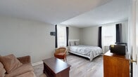 Located at 123 N Broad St in Lancaster, Ohio, this one-bedroom, one-bathroom unit offers a well-organized and efficiently designed living space across a single floor. The interior is finished with light-toned, hardwood-style flooring throughout, lending a clean and cohesive look to the entire unit.

The main living area provides enough room for a sofa, coffee table, and dresser, with a microwave and storage chest offering practical convenience. Natural light filters in through multiple windows dressed with dark curtain panels, brightening the neutral gray walls and creating a comfortable atmosphere.

The bedroom is generously sized relative to the overall footprint of the unit, easily accommodating a full bed, seating, and additional furniture. A wall-mounted TV bracket and a window-unit HVAC system are notable features, along with windows that allow for continued natural light. A textured ceiling adds a subtle architectural detail to this space.

The bathroom is straightforward and functional, finished with beige tile flooring and a tiled walk-in shower. A pedestal sink, standard toilet, and a grab bar reflect a practical, accessible design. The neutral color palette carries throughout, maintaining a consistent and understated aesthetic across all rooms. Overall, the unit presents a tidy, no-frills living environment well-suited for efficient, comfortable everyday use.