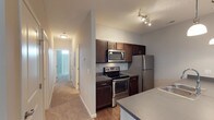 This single-story residence presents a well-organized layout centered around a functional kitchen and a connecting hallway. The kitchen is equipped with a full suite of stainless steel appliances, including a refrigerator, electric range with oven, and an over-the-range microwave. Rich, dark espresso-toned cabinetry lines the upper and lower walls, offering ample storage, while dark countertops provide a cohesive, contemporary aesthetic. A track lighting fixture mounted on the ceiling delivers focused illumination over the cooking area. The countertop extends to form a peninsula, hinting at convenient bar-style seating or additional prep space. Flooring in the kitchen transitions to hardwood-style material, while the adjacent hallway features neutral-toned carpet that continues toward the rest of the unit. The hallway is well-lit with a flush ceiling fixture and leads past multiple doorways, suggesting access to bedrooms and a bathroom. At the far end of the hallway, vertical blinds are visible, indicating a window or door that allows natural light to filter into the corridor. The overall color palette is neutral and understated, creating a clean, move-in-ready atmosphere throughout.