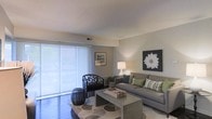 This single-story space centers around a well-appointed living room designed with a neutral, contemporary aesthetic. The room is anchored by large sliding glass doors dressed with sheer white panel curtains, allowing abundant natural light to fill the space while offering views of greenery outside. The soft, diffused light creates a calm and inviting ambiance throughout.

The living area features warm, light-toned walls complemented by dark hardwood-style flooring, establishing a balanced contrast that adds depth to the room. A textured area rug with a circular pattern grounds the seating arrangement, which includes a generously sized gray sofa adorned with green and patterned accent pillows, paired with a distinctive dark lattice-style armchair. A rectangular coffee table serves as a functional centerpiece.

Wall décor includes framed artwork and a bold floral canvas print, adding visual interest without overwhelming the space. A table lamp provides warm ambient lighting, enhancing the room's cozy feel during evening hours. The textured ceiling and visible HVAC vent suggest practical, well-maintained construction. Overall, the space offers a comfortable and stylish living environment.