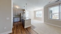 Located in Goose Creek, South Carolina, this single-floor residence offers a thoughtfully designed open-concept layout that seamlessly connects the kitchen, dining, and living areas. The kitchen is well-appointed with gray shaker-style cabinetry, granite countertops, stainless steel appliances including a refrigerator with ice maker, electric range, and over-the-range microwave, as well as a central island with pendant lighting and an integrated sink. Rich hardwood-style flooring runs throughout the main living spaces, transitioning to plush carpet in the living room and bedrooms, creating a warm contrast. The spacious living area features crown molding, a ceiling fan, and direct access to a private balcony through a glass door, offering open views of the surrounding neighborhood. The primary bedroom connects to a well-finished en-suite bathroom showcasing granite-topped vanity cabinetry in a muted blue-gray finish and hardwood-style flooring. A secondary full bathroom mirrors the same quality finishes, complete with a tub-shower combination. A generously sized walk-in closet with multi-tiered wire shelving systems provides abundant storage. Natural light flows throughout via multiple windows with blinds, enhancing the clean, neutral color palette and modern aesthetic of this move-in-ready residence.