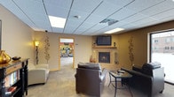 This single-floor amenity space is designed to offer both relaxation and activity in a comfortable, well-appointed setting. The lounge area serves as the centerpiece of the space, featuring a tiled corner fireplace with a wooden mantel and a wall-mounted flat-screen television above it, creating a warm and inviting focal point. Plush leather seating, including a sofa and armchairs, is arranged around a glass-top coffee table, offering a comfortable place to unwind. Decorative metallic wall art with a floral motif adds an elegant touch to the warm, golden-toned walls. The drop-panel ceiling is fitted with recessed lighting panels that provide bright, even illumination throughout. Carpeted flooring in a neutral tone ties the space together seamlessly. A doorway with wood trim leads directly into the fitness center, which is visible from the lounge and features a variety of cardio equipment, including treadmills and elliptical machines, set against vibrant accent walls. The layout promotes a smooth flow between the two areas, making this space a versatile and functional amenity suited for both leisure and wellness activities.