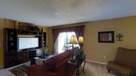 This single-floor space centers around a comfortable and warmly appointed living room designed for relaxed everyday living. The room is finished with a textured ceiling and neutral carpeted flooring, creating a cozy, familiar atmosphere throughout. Walls painted in a warm golden tone give the space an inviting, sunlit feel even on overcast days. A large sliding glass door serves as the focal point of one wall, framed by rich burgundy patterned curtains with an elegant valance that adds a classic decorative touch. The door allows ample natural light to fill the room while offering views to an outdoor area beyond. Built-in shelving and an entertainment unit along one wall provide both functional storage and display space, accommodating a television and decorative accents including plants and collectibles. The seating area features a plush red sofa paired with a dark metal accent table and coordinating floor lamps, lending the room a warm, layered ambiance. A framed piece of wall art adds a finishing touch to the decor. Overall, the space offers a traditional, homey aesthetic with thoughtful details that create a comfortable and welcoming environment.