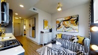 This thoughtfully designed single-floor residence makes a striking first impression with its bold, contemporary aesthetic. The living area serves as the heart of the space, anchored by warm hardwood flooring that runs throughout and bathed in a bright, welcoming ambiance. A large, eye-catching abstract canvas dominates the main wall, showcasing a dynamic palette of yellow, black, mint, and white that sets the tone for the entire interior. The seating area features a patterned sofa adorned with yellow accent pillows, complemented by a graphic black-and-white area rug that adds depth and visual interest underfoot. A sleek, distressed-finish sideboard provides functional storage while doubling as a display surface for decorative accents, including a sculptural vase with yellow florals. A vintage-style pendant light fixture adds a warm, artistic focal point overhead. A hallway leads from the main living area toward additional rooms, with large framed artwork lining the walls and continuing the space's cohesive yellow and black color story. Mirrored closet doors reflect light and enhance the sense of openness. The overall ambiance is vibrant yet curated, blending modern design sensibilities with artistic personality to create a lively and inviting living environment.