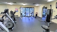 This fitness center is a fully functional workout facility designed to support a wide range of exercise routines. The space features durable rubber flooring throughout, providing safety and comfort during workouts. Large glass doors and windows along the back wall flood the room with natural light and offer clear views of an outdoor patio and landscaped surroundings, creating an open and inviting atmosphere.

The equipment layout is thoughtfully organized to maximize usable floor space. A multi-station cable and weight machine serves as a centerpiece for strength training, accompanied by a dedicated free weights section complete with a full dumbbell rack. A weight bench is positioned nearby for versatile use. Cardio equipment, including what appears to be an elliptical and additional machines, lines the perimeter of the room.

The walls are finished in a warm neutral tone, and recessed fluorescent lighting ensures the space is well-lit at all times. An informational workout poster is displayed on the wall, offering exercise guidance for users. The overall layout is clean, practical, and accommodating for both beginner and experienced fitness enthusiasts, making this a comprehensive amenity space suited for daily use.
