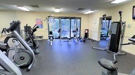 This fitness center is a fully functional workout facility designed to support a wide range of exercise routines. The space features durable rubber flooring throughout, providing safety and comfort during workouts. Large glass doors and windows along the back wall flood the room with natural light and offer clear views of an outdoor patio and landscaped surroundings, creating an open and inviting atmosphere.

The equipment layout is thoughtfully organized to maximize usable floor space. A multi-station cable and weight machine serves as a centerpiece for strength training, accompanied by a dedicated free weights section complete with a full dumbbell rack. A weight bench is positioned nearby for versatile use. Cardio equipment, including what appears to be an elliptical and additional machines, lines the perimeter of the room.

The walls are finished in a warm neutral tone, and recessed fluorescent lighting ensures the space is well-lit at all times. An informational workout poster is displayed on the wall, offering exercise guidance for users. The overall layout is clean, practical, and accommodating for both beginner and experienced fitness enthusiasts, making this a comprehensive amenity space suited for daily use.