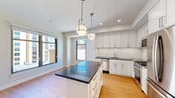 This efficiently designed single-floor unit offers a thoughtful layout that maximizes every square foot. The open-concept kitchen serves as the heart of the space, showcasing crisp white shaker cabinetry, dark stone countertops, and a classic subway tile backsplash. A central kitchen island with a dark countertop provides additional prep space and doubles as a casual dining or gathering surface. Pendant globe lights hang above the island, adding a modern, polished aesthetic. Stainless steel appliances, including a gas range, built-in microwave, dishwasher, and full-size refrigerator, complete the kitchen's functional appeal. Warm hardwood flooring extends throughout the main living area, complementing the clean white walls and creating an inviting ambiance. Large windows allow natural light to flow in and offer views of the surrounding urban streetscape. The bathroom is finished with refined details, including a marble-veined countertop, undermount sink, subway tile walk-in shower, and classic black-and-white mosaic tile flooring. Recessed lighting and a large mirror enhance the sense of brightness and space. Overall, the unit presents a clean, modern aesthetic with quality finishes suited for contemporary urban living.
