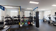 The fitness center at 3050 Plantation Garden Dr in Fayetteville, NC is a well-equipped, single-floor facility designed to support a wide variety of workout routines. The space is organized into distinct training zones, each outfitted with professional-grade equipment. The main floor features a robust functional training rig with TRX suspension straps, a hanging heavy bag for boxing training, and an extensive free weights section with a full dumbbell rack, kettlebells, medicine balls, and plyo boxes. A dedicated cardio area lines the perimeter with multiple treadmills, elliptical machines, stair climbers, and stationary bikes, several equipped with individual monitors. A separate annex room offers additional cardio equipment, including an air bike and elliptical, and features French doors with transom windows that invite natural light and provide views to the exterior. Another adjoining room houses a multi-functional cable machine and an upright bike, finished with clean white walls, crown molding, and rubber flooring throughout. Large skylights and recessed panel lighting keep the entire facility well-illuminated, while full-length mirrors enhance the sense of openness. The rubber flooring across all areas ensures safety and durability, making this fitness center a comprehensive and functional amenity.