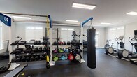 The fitness center at 3050 Plantation Garden Dr in Fayetteville, NC is a well-equipped, single-floor facility designed to support a wide variety of workout routines. The space is organized into distinct training zones, each outfitted with professional-grade equipment. The main floor features a robust functional training rig with TRX suspension straps, a hanging heavy bag for boxing training, and an extensive free weights section with a full dumbbell rack, kettlebells, medicine balls, and plyo boxes. A dedicated cardio area lines the perimeter with multiple treadmills, elliptical machines, stair climbers, and stationary bikes, several equipped with individual monitors. A separate annex room offers additional cardio equipment, including an air bike and elliptical, and features French doors with transom windows that invite natural light and provide views to the exterior. Another adjoining room houses a multi-functional cable machine and an upright bike, finished with clean white walls, crown molding, and rubber flooring throughout. Large skylights and recessed panel lighting keep the entire facility well-illuminated, while full-length mirrors enhance the sense of openness. The rubber flooring across all areas ensures safety and durability, making this fitness center a comprehensive and functional amenity.