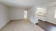 This single-floor space offers a straightforward, functional layout designed for comfortable everyday living. The main living area is finished with light-toned tile flooring that extends throughout, creating a clean and cohesive look that is easy to maintain. Crisp white walls and a standard ceiling height give the space a bright, neutral atmosphere, while a ceiling fan with an integrated light fixture provides both airflow and overhead illumination. A front entry door with a standard latch provides direct access to the living area, which opens toward additional rooms through a wide doorway on the far wall. A partial wall with a built-in upper cabinet element is visible near the entryway, suggesting a transitional area that may connect to a kitchen or dining space. Through the open doorway, a glimpse of a darkly finished cabinet hints at a kitchen beyond. The layout flows naturally from the entry through the main living space and into the rest of the unit, making efficient use of the available floor plan. The overall ambiance is clean, minimal, and move-in ready, offering a blank canvas that is well-suited for a variety of furnishing styles and personal touches.