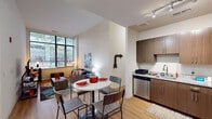 This single-floor residence offers a thoughtfully designed open-concept layout that seamlessly connects the kitchen, dining, and living areas. The kitchen is outfitted with warm wood-toned cabinetry, granite countertops, and a full suite of stainless steel appliances, including a refrigerator, electric range, microwave, and dishwasher. Track lighting overhead provides ample illumination. Adjacent to the kitchen, the dining area accommodates a round table with seating for four, while the living space beyond benefits from expansive floor-to-ceiling windows that invite generous natural light and offer views of surrounding greenery. Light hardwood flooring runs throughout the main living areas, contributing to a bright and cohesive aesthetic. The residence includes multiple bedrooms, each featuring large windows with outdoor views, creating airy and comfortable retreats. The bathrooms are well-appointed with granite-topped wood vanities, full soaking tubs with shower combinations, and hardwood-style flooring. Clean white walls and neutral tones throughout the unit create a consistent, modern atmosphere that is both functional and inviting.