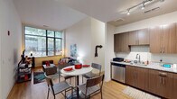 This single-floor residence offers a thoughtfully designed open-concept layout that seamlessly connects the kitchen, dining, and living areas. The kitchen is outfitted with warm wood-toned cabinetry, granite countertops, and a full suite of stainless steel appliances, including a refrigerator, electric range, microwave, and dishwasher. Track lighting overhead provides ample illumination. Adjacent to the kitchen, the dining area accommodates a round table with seating for four, while the living space beyond benefits from expansive floor-to-ceiling windows that invite generous natural light and offer views of surrounding greenery. Light hardwood flooring runs throughout the main living areas, contributing to a bright and cohesive aesthetic. The residence includes multiple bedrooms, each featuring large windows with outdoor views, creating airy and comfortable retreats. The bathrooms are well-appointed with granite-topped wood vanities, full soaking tubs with shower combinations, and hardwood-style flooring. Clean white walls and neutral tones throughout the unit create a consistent, modern atmosphere that is both functional and inviting.