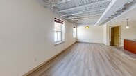 Located at 4001 E Genesee St in Syracuse, NY, this two-bedroom, two-bathroom residence offers a distinctive loft-inspired aesthetic defined by its striking exposed steel truss ceilings that lend an industrial character throughout the single-floor layout. The open-concept kitchen and living area serves as the heart of the home, anchored by a central island topped with dark granite countertops, a stainless steel sink, and pendant lighting overhead. Rich wood cabinetry lines the kitchen walls alongside a full suite of stainless steel appliances, including a refrigerator, electric range, and microwave. Light hardwood flooring flows through the living and bedroom areas, complementing the bright, neutral-toned walls. The generously sized primary bedroom benefits from multiple windows that invite natural light and offer views of the surrounding wooded landscape, while the exposed truss ceiling adds volume and visual interest. The second bedroom is equally well-lit and spacious. Both bathrooms are finished with tile flooring and warm wood vanities; one features a walk-in tiled shower, while the other includes an arched mirror and stylish lighting fixture. An in-unit laundry room adds everyday convenience to this unique and characterful residence.