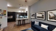This single-floor space showcases a thoughtfully designed open-concept layout that seamlessly connects the kitchen and living area, creating a cohesive and inviting atmosphere. The kitchen is a standout feature, outfitted with dark espresso cabinetry, stainless steel appliances, a mosaic tile backsplash, and light stone countertops. A peninsula-style breakfast bar with bar stool seating adds both functionality and a casual dining option. Overhead, a series of pendant lights provide warm, focused illumination that complements the kitchen's contemporary aesthetic. The living area benefits from soaring ceilings that amplify the sense of space, while warm hardwood-style flooring runs throughout, adding a touch of natural warmth to the cool, neutral color palette. Soft gray walls serve as a versatile backdrop, highlighted by bold framed wall art that adds personality and visual interest. A ceiling fan overhead ensures year-round comfort. The overall ambiance is modern and urban, striking a balance between stylish design and everyday livability — ideal for those who appreciate clean lines, quality finishes, and an open, airy feel.