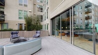 This outdoor courtyard amenity space is set within a contemporary multi-story residential building complex, offering a well-appointed environment for relaxation and socializing. The courtyard is paved with neutral-toned interlocking stone pavers that lend a clean, modern aesthetic throughout.

At the heart of the space, a central fire pit with a glass-bowl design serves as a focal point, surrounded by stylish blue lounge chairs and a curved sectional sofa — ideal for gathering. A freestanding egg-shaped hanging chair adds a casual, playful element to one corner of the courtyard.

Raised concrete planter beds line the perimeter, filled with mature bamboo and ornamental trees that provide a sense of privacy and natural greenery against the building's neutral facade. A secondary courtyard area extends further, featuring a raised circular garden bed with low ornamental plantings and bistro-style seating, flanked by suspended hanging chairs and curved white seating.

Floor-to-ceiling sliding glass doors connect the outdoor space directly to an interior lounge, which is visible through the glazing and features warm pendant lighting, booth seating, and dining tables — creating a seamless indoor-outdoor flow. The surrounding building architecture, with its balconies and large windows, frames the courtyard and contributes to a sense of enclosure and urban sophistication.