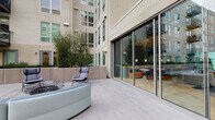 This outdoor courtyard amenity space is set within a contemporary multi-story residential building complex, offering a well-appointed environment for relaxation and socializing. The courtyard is paved with neutral-toned interlocking stone pavers that lend a clean, modern aesthetic throughout.

At the heart of the space, a central fire pit with a glass-bowl design serves as a focal point, surrounded by stylish blue lounge chairs and a curved sectional sofa — ideal for gathering. A freestanding egg-shaped hanging chair adds a casual, playful element to one corner of the courtyard.

Raised concrete planter beds line the perimeter, filled with mature bamboo and ornamental trees that provide a sense of privacy and natural greenery against the building's neutral facade. A secondary courtyard area extends further, featuring a raised circular garden bed with low ornamental plantings and bistro-style seating, flanked by suspended hanging chairs and curved white seating.

Floor-to-ceiling sliding glass doors connect the outdoor space directly to an interior lounge, which is visible through the glazing and features warm pendant lighting, booth seating, and dining tables — creating a seamless indoor-outdoor flow. The surrounding building architecture, with its balconies and large windows, frames the courtyard and contributes to a sense of enclosure and urban sophistication.