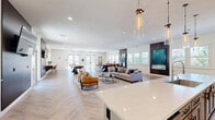 This single-floor clubhouse at 500 Roseville Pkwy in Roseville, CA, offers a generously sized, open-concept layout that seamlessly blends a fully equipped kitchen with a comfortable living and recreation area. The space is unified by striking herringbone-patterned light wood flooring throughout, lending a contemporary and polished aesthetic.

The kitchen is a standout feature, showcasing soft gray shaker cabinetry, a textured tile backsplash, open display shelving, and glass-front upper cabinets. A large center island with a crisp white quartz countertop provides ample bar seating with sleek black leather stools, while pendant lights with clear glass cylinders hang overhead. A professional-grade faucet and stainless steel sink anchor the island, and a full-size stainless steel refrigerator completes the space.

The adjacent living area is bright and welcoming, filled with natural light from multiple windows with plantation shutters. Dark wood accent walls serve as a focal point, each mounting a flat-screen television. Comfortable cognac leather lounge chairs and a cozy sofa create inviting gathering spaces. A shuffleboard table and pool table add recreational appeal, while a wall-mounted cue rack and a counter-height bar ledge along the windows provide additional seating and functionality. The overall ambiance is modern, warm, and well-appointed.