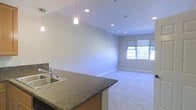 This single-floor space showcases a thoughtfully designed open-concept layout that seamlessly connects the kitchen and living areas. The kitchen is appointed with a dark granite countertop island that doubles as a breakfast bar, a stainless steel double sink, and warm wood cabinetry. Stylish pendant lights hang above the island, complementing a series of recessed ceiling lights that illuminate the space evenly throughout.

The living area flows naturally from the kitchen, finished with plush neutral-toned carpeting that adds warmth underfoot. Light gray walls provide a clean, versatile backdrop, while a large window brings in natural light and offers a view of the surrounding greenery outside. The window is fitted with a simple roller shade for privacy and light control.

The ceilings are smooth and well-proportioned, contributing to an airy, uncluttered ambiance. The overall design emphasizes an open, functional floor plan that makes efficient use of space, with a neutral color palette throughout that lends a modern yet welcoming feel. The combination of natural light, quality finishes, and open sightlines makes this a comfortable and practical single-level space.