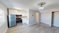 This single-floor space offers a clean, modern aesthetic with thoughtful finishes throughout. The kitchen is a standout feature, showcasing white shaker-style cabinetry that extends to the upper walls, providing generous storage. A mosaic tile backsplash adds a touch of texture and visual interest, complementing the light-toned countertops. The kitchen is equipped with a full suite of stainless steel appliances, including a refrigerator, an electric range with a smooth cooktop, and an over-the-range microwave, making it both functional and contemporary in appearance. Light hardwood-style flooring runs throughout the space, lending warmth and continuity between areas. The open layout connects the kitchen to the adjacent living area, which is illuminated by a ceiling fan with an integrated light fixture, enhancing airflow and brightness. Neutral grey walls and crisp white trim create a fresh, versatile backdrop suitable for a variety of interior styles. A textured ceiling adds a classic touch, while a hallway visible from the main living area suggests additional rooms beyond. Overall, the space presents a well-maintained, move-in-ready environment with a cohesive and practical design.
