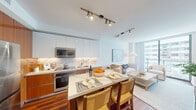 This single-floor residence is thoughtfully designed with a contemporary aesthetic, seamlessly blending function and style across its well-appointed spaces. Rich hardwood flooring flows throughout, providing a warm contrast to the crisp white walls and ceilings.

The open-plan kitchen and living area serves as the heart of the home. The kitchen features a dual-tone design, combining white gloss upper cabinetry with warm wood-grain lower cabinets, complemented by marble-effect backsplash, stainless steel appliances, and a waterfall-edge island with seating. Track lighting illuminates the space effectively.

The living area transitions naturally from the kitchen, offering floor-to-ceiling sliding glass doors that open to a private balcony, allowing generous natural light and outward views of the surrounding cityscape. A coastal-inspired color palette of blues and neutrals reinforces a relaxed ambiance.

The bedroom is a calm retreat, featuring a ceiling fan, a soft blue accent wall, a dedicated workspace, a walk-in closet, and direct access to the ensuite bathroom. The bathroom is finished with marble-effect tiling on both floors and walls, a glass-enclosed shower, and a wood-trimmed vanity with a white stone countertop.