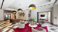 This single-floor amenity space showcases a sophisticated contemporary design that seamlessly blends bold aesthetics with functional comfort. The entrance features large floor-to-ceiling windows and folding glass doors that flood the interior with natural light and offer views of the surrounding streetscape. A striking geometric patterned tile floor in tones of charcoal, white, and beige flows throughout, anchoring the space with visual dynamism.

The main lounge area is centered around a vibrant crimson area rug, atop which plush gray velvet armchairs are arranged around a sleek marble-topped coffee table. Overhead, dramatic matte black dome pendant lights with gold interiors provide warm, focused illumination. A decorative wall installation of clustered metallic disc sculptures serves as a distinctive artistic focal point.

Adjacent to the lounge is a large communal dining table surrounded by woven-back wood chairs, ideal for collaborative gatherings or shared meals. The space is further enhanced by a dedicated kitchenette featuring gray cabinetry, blush-toned subway tile backsplash, and a built-in wine cooler — all lit by a sleek track lighting system. A secondary lounge area with bar-height seating and a wall-mounted television offers additional flexible gathering space, making this a well-appointed and versatile communal environment.