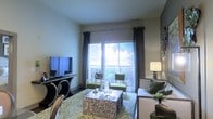 This single-story space showcases a thoughtfully designed living room that serves as the focal point of the interior. Large sliding glass doors dominate one wall, allowing abundant natural light to fill the room while providing direct access to a private balcony overlooking lush greenery. Vertical blinds and floor-length charcoal drape panels frame the doors, offering both style and light control.

The living area is furnished with a cohesive contemporary arrangement, including a plush sofa accented with decorative pillows, two green accent chairs, and a mosaic-tiled coffee table that adds a distinctive visual element. A patterned area rug in soft greens and neutrals anchors the seating arrangement, complementing the overall color palette.

An elegant console table with curved metal legs supports a flat-screen television on one side of the room, while a stylish table lamp provides warm ambient lighting on the other. The smooth, neutral-toned walls and clean ceiling contribute to an open, airy atmosphere. Together, the furnishings, natural light, and outdoor access create a comfortable and visually appealing living environment.