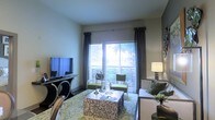 This single-story space showcases a thoughtfully designed living room that serves as the focal point of the interior. Large sliding glass doors dominate one wall, allowing abundant natural light to fill the room while providing direct access to a private balcony overlooking lush greenery. Vertical blinds and floor-length charcoal drape panels frame the doors, offering both style and light control.

The living area is furnished with a cohesive contemporary arrangement, including a plush sofa accented with decorative pillows, two green accent chairs, and a mosaic-tiled coffee table that adds a distinctive visual element. A patterned area rug in soft greens and neutrals anchors the seating arrangement, complementing the overall color palette.

An elegant console table with curved metal legs supports a flat-screen television on one side of the room, while a stylish table lamp provides warm ambient lighting on the other. The smooth, neutral-toned walls and clean ceiling contribute to an open, airy atmosphere. Together, the furnishings, natural light, and outdoor access create a comfortable and visually appealing living environment.