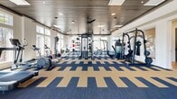 Located at 2920 Frontage Rd in Gainesville, GA, this fitness center offers a generously sized, single-floor exercise room designed to support a comprehensive workout experience. The space features a striking slatted wood ceiling accented by circular ring pendant lights and ceiling fans, lending a modern yet warm aesthetic throughout. Underfoot, a bold navy and tan geometric carpet tile pattern adds visual energy to the open floor plan.

Large multi-pane windows line the walls, flooding the interior with abundant natural light and offering serene views of the surrounding wooded landscape. The spacious layout accommodates a wide array of equipment, including multiple treadmills, rowing machines, stationary bikes, cable machines, functional trainers, weight racks with dumbbells, adjustable benches, and strength machines. A large wall-mounted flat-screen TV serves as a central entertainment focal point, enhancing the workout environment.

Mirrors are strategically placed to amplify the sense of space and allow users to monitor their form. The well-organized arrangement of equipment ensures clear circulation paths throughout the room. Ceiling fans and recessed lighting supplement the natural light, maintaining a comfortable atmosphere. This fitness center delivers a polished, fully equipped environment suited for a wide range of fitness routines and training styles.