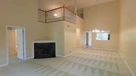 This two-story home makes an immediate impression with its dramatic vaulted ceilings that rise through both floors, creating a grand sense of openness in the main living area. The centerpiece of the living room is a classic fireplace framed in crisp white with a dark surround, offering both a functional and aesthetic focal point. Above, a wood-railed second-floor balcony overlooks the main living space, adding architectural depth and an elegant sense of connectivity between floors. Neutral, warm-toned walls complement the patterned carpeting throughout, lending the home a cohesive and inviting palette. An elegant chandelier anchors the adjacent dining or secondary living area, which benefits from natural light streaming in through a window with views of greenery outside. Wide open sightlines flow seamlessly from room to room, emphasizing the home's spacious and airy layout. A hallway visible from the main living area hints at additional rooms beyond, while a dedicated entryway provides a clear transition into the home. The overall ambiance is bright, open, and welcoming, with traditional architectural details that add character and charm throughout.