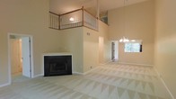 This two-story home makes an immediate impression with its dramatic vaulted ceilings that rise through both floors, creating a grand sense of openness in the main living area. The centerpiece of the living room is a classic fireplace framed in crisp white with a dark surround, offering both a functional and aesthetic focal point. Above, a wood-railed second-floor balcony overlooks the main living space, adding architectural depth and an elegant sense of connectivity between floors. Neutral, warm-toned walls complement the patterned carpeting throughout, lending the home a cohesive and inviting palette. An elegant chandelier anchors the adjacent dining or secondary living area, which benefits from natural light streaming in through a window with views of greenery outside. Wide open sightlines flow seamlessly from room to room, emphasizing the home's spacious and airy layout. A hallway visible from the main living area hints at additional rooms beyond, while a dedicated entryway provides a clear transition into the home. The overall ambiance is bright, open, and welcoming, with traditional architectural details that add character and charm throughout.