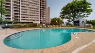 Located at 12540 Edgewater Dr in Lakewood, Ohio, this outdoor pool area serves as a premier recreational amenity for the high-rise residential community situated along Lake Erie's shoreline. The centerpiece of the space is a generously proportioned freeform swimming pool with a smooth, curved design that accommodates both lap swimming and leisurely use. The pool features stainless steel entry rails at multiple points for easy access, and its clear turquoise water reflects the open sky above.

Surrounding the pool is a broad, textured concrete sun deck that provides ample room for lounging and movement. The deck is furnished with multiple green reclining lounge chairs positioned along the perimeter, along with a shaded seating area featuring a blue umbrella and patio table — ideal for relaxing out of direct sunlight. The entire pool area is enclosed by a black iron perimeter fence with a gated entry, ensuring both security and privacy.

Mature trees line the edges of the space, providing natural shade and greenery. The backdrop features the community's towering residential high-rise buildings, which rise prominently and are visible from multiple vantage points within the pool area. Open sightlines toward the surrounding landscape hint at the proximity to Lake Erie. A small storage shed near the gate provides convenient poolside storage for equipment and supplies.