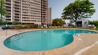 Located at 12540 Edgewater Dr in Lakewood, Ohio, this outdoor pool area serves as a premier recreational amenity for the high-rise residential community situated along Lake Erie's shoreline. The centerpiece of the space is a generously proportioned freeform swimming pool with a smooth, curved design that accommodates both lap swimming and leisurely use. The pool features stainless steel entry rails at multiple points for easy access, and its clear turquoise water reflects the open sky above.

Surrounding the pool is a broad, textured concrete sun deck that provides ample room for lounging and movement. The deck is furnished with multiple green reclining lounge chairs positioned along the perimeter, along with a shaded seating area featuring a blue umbrella and patio table — ideal for relaxing out of direct sunlight. The entire pool area is enclosed by a black iron perimeter fence with a gated entry, ensuring both security and privacy.

Mature trees line the edges of the space, providing natural shade and greenery. The backdrop features the community's towering residential high-rise buildings, which rise prominently and are visible from multiple vantage points within the pool area. Open sightlines toward the surrounding landscape hint at the proximity to Lake Erie. A small storage shed near the gate provides convenient poolside storage for equipment and supplies.