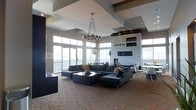 This single-floor space showcases a refined, contemporary design that immediately captures attention with its dramatic architectural ceiling details — a series of recessed and layered geometric panels that add depth and visual interest overhead. Two striking modern chandeliers with spherical metallic accents serve as bold focal points, casting a warm, ambient glow throughout the open interior.

The expansive open-concept layout is anchored by a large, plush sectional sofa in deep charcoal, paired with a sleek, multi-tiered dark coffee table atop a richly patterned carpet in warm earth tones. Adjacent to the lounge area, a sophisticated dining space is outfitted with contemporary white chairs and clean-lined tables, ideal for gathering and entertaining.

A floor-to-ceiling stone fireplace commands one wall, flanked by built-in display niches, lending the space a sense of grandeur and warmth. Generous windows wrap around the perimeter, flooding the interior with natural light and offering expansive views of the surrounding exterior. The overall ambiance is polished and upscale, blending neutral tones of gray, white, and warm brown to create an atmosphere that is both inviting and impressively designed.