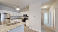 This single-story residence offers a thoughtfully organized layout across two bedrooms and two bathrooms. The open-concept kitchen serves as a central highlight, featuring crisp white cabinetry, granite countertops with subtle veining, and a full suite of stainless steel appliances including a range, over-the-range microwave, refrigerator, and dishwasher. A peninsula with a undermount sink and pendant lighting above enhances both functionality and visual appeal. Light wood-style flooring runs throughout the kitchen and entry areas, lending a clean, modern aesthetic.

The living room connects seamlessly to the main living area, finished with neutral carpet and illuminated by natural light through French doors. The entry area transitions between the hardwood-style flooring and carpet, clearly delineating spaces. Both bedrooms are carpeted and feature windows with blinds that allow for controlled natural light. The primary bedroom is generously sized and includes a walk-in closet outfitted with wire shelving systems. The bathrooms include a walk-in shower with built-in corner shelving. Neutral wall tones throughout the residence provide a cohesive, move-in-ready ambiance.