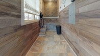 This dedicated pet spa is a well-appointed, single-level grooming facility designed with both functionality and style in mind. The space is finished with durable, large-format stone tile flooring in warm earth tones that flows throughout the entire area, making cleanup effortless. The lower half of the walls is clad in wood-look tile paneling, creating a warm, welcoming aesthetic while remaining practical and moisture-resistant.

The focal point of the space is a built-in pet bathing station, featuring a raised, tiled wash basin with step access, a wall-mounted handheld spray nozzle, and a dedicated drying hose — all thoughtfully integrated into the tiled surround. The backsplash area of the bathing station showcases a diagonal-set decorative tile pattern, adding a refined design detail. Playful paw print wall decals add a charming, pet-friendly touch throughout the upper walls.

Natural light fills the space through a slatted-blind window adjacent to the bathing area, while a glass-panel entry door at the far end of the room provides additional brightness and a clear view to the exterior. The entry corridor is spacious and open, offering ample room for maneuvering pets. An electrical panel is neatly mounted to the wall, and a coat hook near the entrance adds a practical finishing touch to this well-designed pet care space.