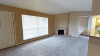 This single-floor space offers a welcoming and functional layout centered around a generously sized living area. The room is finished with soft, neutral-toned wall-to-wall carpeting that adds warmth underfoot, complemented by warm beige walls that create a comfortable, inviting atmosphere throughout.

A standout architectural feature is the vaulted ceiling, which angles upward to lend the living space a sense of openness and volume. Natural light flows freely through a large picture window fitted with horizontal blinds, allowing for adjustable light control while maintaining a bright and airy feel.

In the corner of the living area sits a classic brick fireplace, serving as a natural focal point. Its traditional design adds character and charm, making it ideal for creating a cozy gathering space. The fireplace surround and hearth are finished in light-toned brick, consistent with the room's overall neutral palette.

White baseboards and clean wall lines contribute to the tidy, well-maintained appearance of the space. The open, uncluttered floor plan allows for versatile furniture arrangements and makes the living area feel expansive. Overall, the space combines practical design with warm, understated aesthetics, making it a comfortable and adaptable environment.