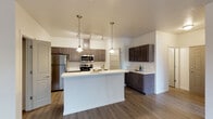 This single-floor residence offers a thoughtfully designed open-concept layout that seamlessly connects the kitchen and living areas. The kitchen is a standout feature, showcasing dark espresso-toned cabinetry complemented by lighter upper cabinets, bright white countertops, and a full suite of stainless steel appliances including a refrigerator, electric range, and built-in microwave. A spacious kitchen island with an integrated sink provides both additional prep space and a natural gathering point. Pendant lighting and a track lighting system overhead add both function and style to the cooking space. A dedicated walk-in pantry offers generous storage directly off the kitchen. Wood-style plank flooring runs throughout the main living areas, lending a warm, cohesive feel, while soft carpet adds comfort to the bedroom, which includes a closet with built-in shelving. The bathroom features a combination tub and shower, a single-sink vanity with dark cabinetry, and wood-style flooring that ties the space together. A dedicated laundry room houses a full-size washer and dryer with wire shelving above for added storage. The open living area is generously sized, filled with natural light, and provides a versatile, move-in-ready space throughout.