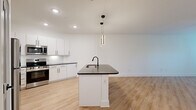 This well-designed single-floor residence offers a thoughtful layout across one bedroom, one bathroom, a closet, and a combined kitchen and living area connected by a hallway. The open-concept kitchen and living room serve as the heart of the home, featuring light wood-tone flooring that extends seamlessly throughout the entire unit. The kitchen is appointed with crisp white shaker cabinetry, dark countertops, a subway tile backsplash, stainless steel appliances including a range, microwave, and refrigerator, and a central island with a dark countertop, undermount sink, and matte black faucet. Stylish pendant lighting with a modern cylindrical design hangs above the island, adding a contemporary accent. The living area is bright and airy, benefiting from natural light through large windows and a glass-panel entry door, and is equipped with a ceiling fan for added comfort. The bedroom is a calm, well-proportioned retreat with the same warm flooring and a ceiling fan. The bathroom features a clean, minimalist design with a soaking tub and shower combo, matte black fixtures, white wall surround, and light flooring that ties in with the rest of the unit. The overall aesthetic is modern and cohesive, with white walls and black hardware accents creating a clean, contemporary ambiance.