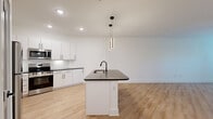 This well-designed single-floor residence offers a thoughtful layout across one bedroom, one bathroom, a closet, and a combined kitchen and living area connected by a hallway. The open-concept kitchen and living room serve as the heart of the home, featuring light wood-tone flooring that extends seamlessly throughout the entire unit. The kitchen is appointed with crisp white shaker cabinetry, dark countertops, a subway tile backsplash, stainless steel appliances including a range, microwave, and refrigerator, and a central island with a dark countertop, undermount sink, and matte black faucet. Stylish pendant lighting with a modern cylindrical design hangs above the island, adding a contemporary accent. The living area is bright and airy, benefiting from natural light through large windows and a glass-panel entry door, and is equipped with a ceiling fan for added comfort. The bedroom is a calm, well-proportioned retreat with the same warm flooring and a ceiling fan. The bathroom features a clean, minimalist design with a soaking tub and shower combo, matte black fixtures, white wall surround, and light flooring that ties in with the rest of the unit. The overall aesthetic is modern and cohesive, with white walls and black hardware accents creating a clean, contemporary ambiance.