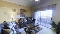 This single-floor residence offers a thoughtfully designed living space that balances comfort and style. The living room serves as the heart of the home, featuring plush, dark upholstered sofas arranged around a glass-top coffee table, creating an inviting area for relaxation and entertaining. A colorful area rug anchors the seating arrangement, adding warmth and visual interest to the space. Crown molding details along the ceiling lend a polished, refined touch to the room, while a ceiling fan ensures year-round comfort. Decorative wall art adds personality and character to the neutral-toned walls. A standout feature of the living area is the large sliding glass door, which floods the room with natural light and opens directly onto a private balcony. The balcony is outfitted with outdoor seating, offering a pleasant spot to enjoy the surrounding views. Sheer curtains and valances frame the glass door, softening the transition between indoor and outdoor living. The overall ambiance is warm and residential, with a layout that makes efficient use of the available space while maintaining an open, airy feel throughout.