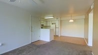 This single-floor space showcases an open-concept layout that seamlessly connects the living area and kitchen. Upon entry, a standard front door with a deadbolt opens into a carpeted living space with neutral walls and a simple, clean aesthetic. The flooring transitions from carpet in the living area to hard flooring in the kitchen zone, clearly delineating the two spaces.

The kitchen features white cabinetry lining the upper and lower sections, a white refrigerator, and a peninsula-style counter that partially separates the kitchen from the living area. A flush-mounted fluorescent ceiling light illuminates the kitchen workspace, while a pendant light hangs over the transitional dining or gathering area, adding a touch of visual interest.

The living area is generously sized with light-toned carpet and plain white walls that lend a bright, neutral backdrop. The overall ambiance is understated and functional, with a practical layout well-suited for everyday living. The space benefits from a straightforward, no-frills design that provides a versatile foundation for personalization.
