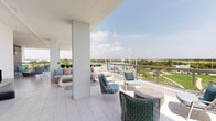 This generously proportioned terrace is designed to function as a complete outdoor living environment, seamlessly blending relaxation, dining, and entertaining. Finished throughout in large-format light-toned tiles, the space is organized into distinct yet flowing zones separated by structural white columns that lend an architectural rhythm without interrupting the openness of the layout.

The lounge area features curved, wicker-base sofas adorned with soft teal and blush cushions, complemented by woven accent chairs in coordinating tones. A patterned area rug anchors this seating zone, adding texture and visual interest underfoot. Adjacent to it, a dedicated dining zone centers around a sleek dark table and woven rope chairs, set atop a boldly graphic medallion rug.

The standout feature of the dining area is a full-height accent wall clad in deep teal three-dimensional tiles, which serves as a dramatic backdrop for a wall-mounted television and a stainless steel outdoor kitchen complete with cabinetry, a sink, and under-counter refrigeration. A globe pendant light with a brass fixture illuminates this zone with warm, ambient light.

The terrace opens entirely to the outdoors, offering expansive panoramic views of manicured green space, treetops, and the surrounding cityscape beneath open skies.