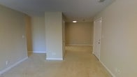 This single-floor space presents a clean, functional layout characterized by soft neutral wall tones and wall-to-wall carpeting throughout. The flooring is finished in a light beige carpet that extends seamlessly across the open areas, contributing to a cohesive and understated aesthetic.

The main area is defined by a partial wall or structural column that subtly divides the space while maintaining an open feel. This architectural element creates a natural sense of separation between zones without fully enclosing them, allowing for flexible use of the space.

Recessed and flush-mounted ceiling lights provide interior illumination, complementing the textured popcorn ceiling finish. White baseboards line the perimeter of each room, offering a crisp contrast against the neutral walls.

A white paneled door is visible toward the right side of the space, indicating access to an additional room or exit point. The overall ambiance is quiet and neutral, offering a blank canvas adaptable to various configurations. The layout appears straightforward, with clearly defined pathways connecting the distinct areas of the interior.