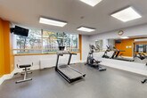Located at 65 Wynford Heights Cres in Toronto, this fitness facility is organized across two connected gym areas, offering a comprehensive workout environment. The main cardio zone features a treadmill, elliptical trainer, and rowing machine, all positioned to take advantage of large windows that frame lush, tree-lined outdoor views — creating a bright and motivating atmosphere. A full-length wall mirror enhances the sense of space while allowing users to monitor their form during exercise. Durable speckled rubber flooring runs throughout, providing both safety and comfort underfoot. Recessed panel lighting illuminates the space evenly from above. The secondary workout area expands the facility's capabilities with a multi-functional cable machine positioned beside additional windows overlooking greenery, a stationary bike, push-up bars, an exercise mat, and a wall-mounted display screen. Bold orange accent walls add an energetic visual contrast to the otherwise clean white interior. A wall-mounted air conditioning unit ensures climate comfort year-round, and a water fountain is conveniently available. Together, the two areas provide a well-rounded space suitable for cardio training, strength work, and functional fitness exercises.