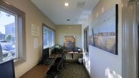 This single-floor office space offers a narrow, elongated layout designed to maximize functionality within a modest footprint. The room features neutral-toned walls complemented by warm, carpeted flooring in an earthy, patterned design that adds texture to the space.

Along one side, a built-in wooden desk runs the length of the wall, providing ample workspace equipped with desktop computers, keyboards, and office essentials—well-suited for administrative or business operations. Ergonomic office chairs accompany the workstations, reinforcing the space's professional purpose.

Recessed ceiling lights provide consistent overhead illumination, supplemented by natural light entering through a window on the adjacent wall, keeping the space bright and practical. HVAC vents integrated into the ceiling ensure climate control throughout.

A notable decorative element is the large landscape mural adorning one wall, adding visual interest and an artistic touch to an otherwise utilitarian environment. A decorative archway-style display piece positioned at the far end of the room serves as a focal point. Posted notices and informational sheets are displayed near the entrance, suggesting a shared or public-facing workspace.