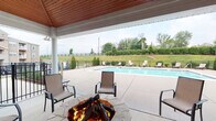 Located at 4121 Reserve Rd in Lexington, Kentucky, this outdoor amenity space is a well-appointed recreational area designed for relaxation and socializing. The centerpiece of the space is a sparkling swimming pool enclosed by a sleek black iron fence, surrounded by a generous concrete deck lined with sling lounge chairs — ideal for sunbathing or unwinding outdoors. A covered pavilion anchors one side of the pool area, featuring a vaulted ceiling with warm wood paneling and a ceiling fan for comfortable use during warm weather. The pavilion is built with classic brick construction and opens via white-framed French doors, offering a seamless transition between indoor and outdoor areas. A prominent stone fire pit surrounded by sling patio chairs serves as a natural gathering focal point beneath the covered structure. The surrounding landscape features manicured hedgerows, ornamental grasses, and mature trees, providing a lush green backdrop. The view from the patio looks out to rolling open land with black wooden fencing characteristic of the Kentucky Bluegrass region. The broader community building is visible in the background, reflecting a traditional multi-story brick architectural style that complements the amenity area's design.